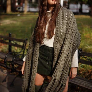 May include: A woman wearing a green crochet shawl with fringe. The shawl has a white crocheted border. She is wearing a white turtleneck sweater and a dark green skirt.