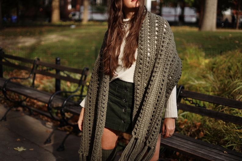 May include: A woman wearing a green crochet shawl with fringe. The shawl has a white crocheted border. She is wearing a white turtleneck sweater and a dark green skirt.