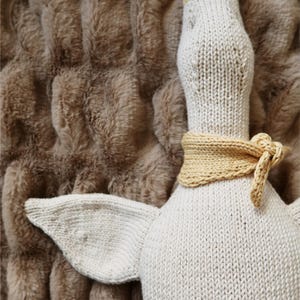 May include: A knitted goose toy in cream and tan. The goose has a cream-colored body, a tan beak and head, and a tan scarf tied around its neck. The goose is lying on a brown, textured surface.