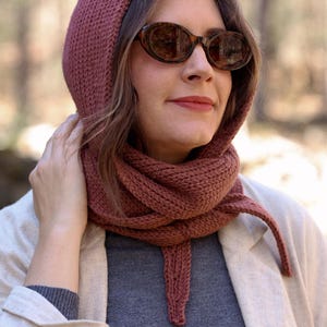 K&ouml;nnte beinhalten: Nahaufnahme einer Frau, die einen rostigen, gestrickten Kapuzen-Schal mit langem Band tr&auml;gt. Der Schal wird &uuml;ber einem grauen Rollkragenpullover und einer cremefarbenen Jacke getragen. Die Frau tr&auml;gt ovale Sonnenbrillen.