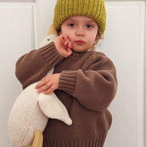 May include: A child wearing a mustard yellow knit beanie and a brown sweater, holding a large white and beige knitted goose toy. The goose has yellow feet. The child is looking at the camera.