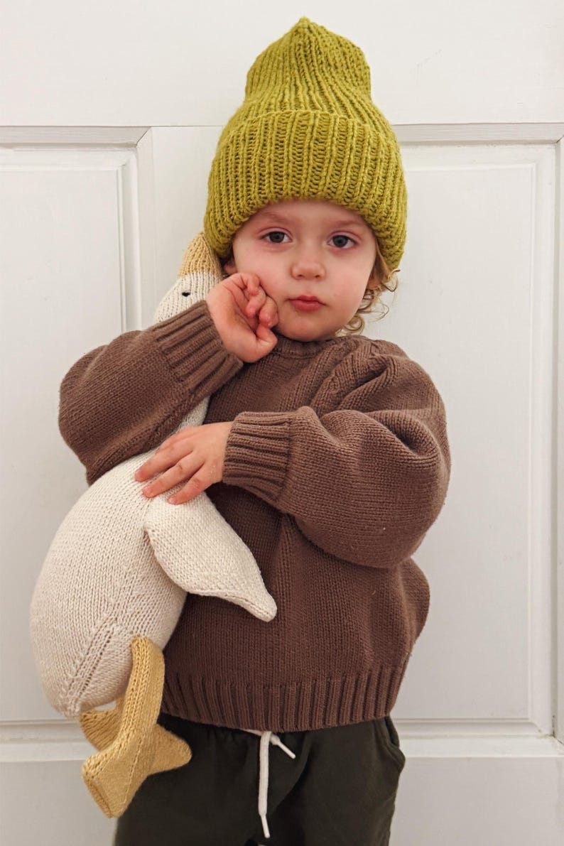 May include: A child wearing a mustard yellow knit beanie and a brown sweater, holding a large white and beige knitted goose toy. The goose has yellow feet. The child is looking at the camera.