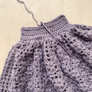 May include: A close-up of a purple crocheted garment with a crochet hook in the top edge.