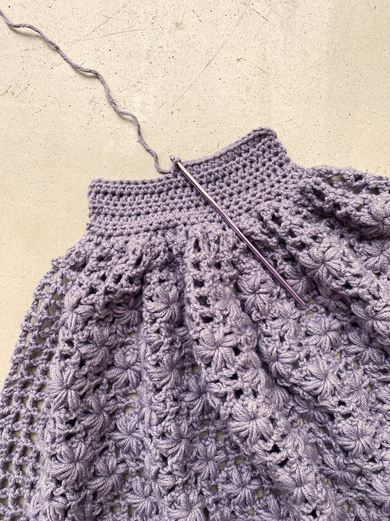 May include: A close-up of a purple crocheted garment with a crochet hook in the top edge.
