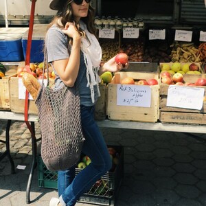 May include: A woman wearing a grey t-shirt, a white scarf, a brown hat, and blue jeans is holding a red apple. She is standing in front of a table with a variety of apples in wooden crates. The crates are labeled with prices and the types of apples. The text on the crates reads "Red Delicious Apples $2.75/lb" and "Empire $2.75/lb".