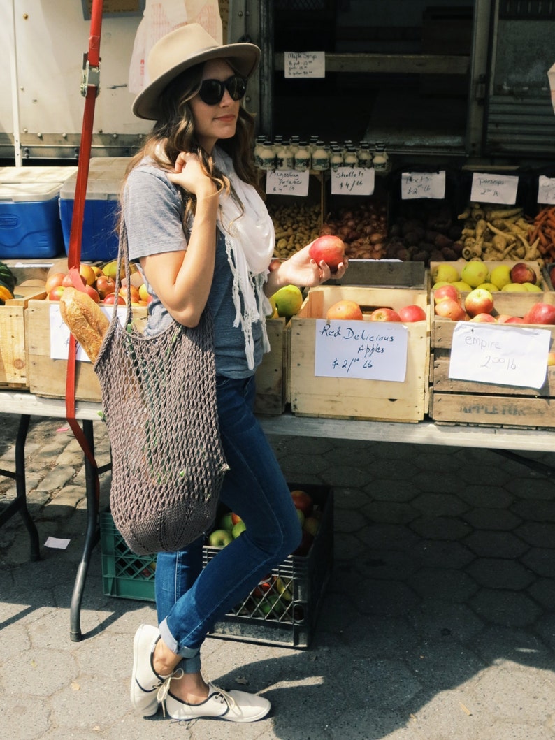 May include: A woman wearing a grey t-shirt, a white scarf, a brown hat, and blue jeans is holding a red apple. She is standing in front of a table with a variety of apples in wooden crates. The crates are labeled with prices and the types of apples. The text on the crates reads "Red Delicious Apples $2.75/lb" and "Empire $2.75/lb".