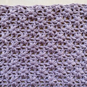 May include: A close-up of a crocheted fabric with a repeating pattern of small flowers. The fabric is a light purple color.