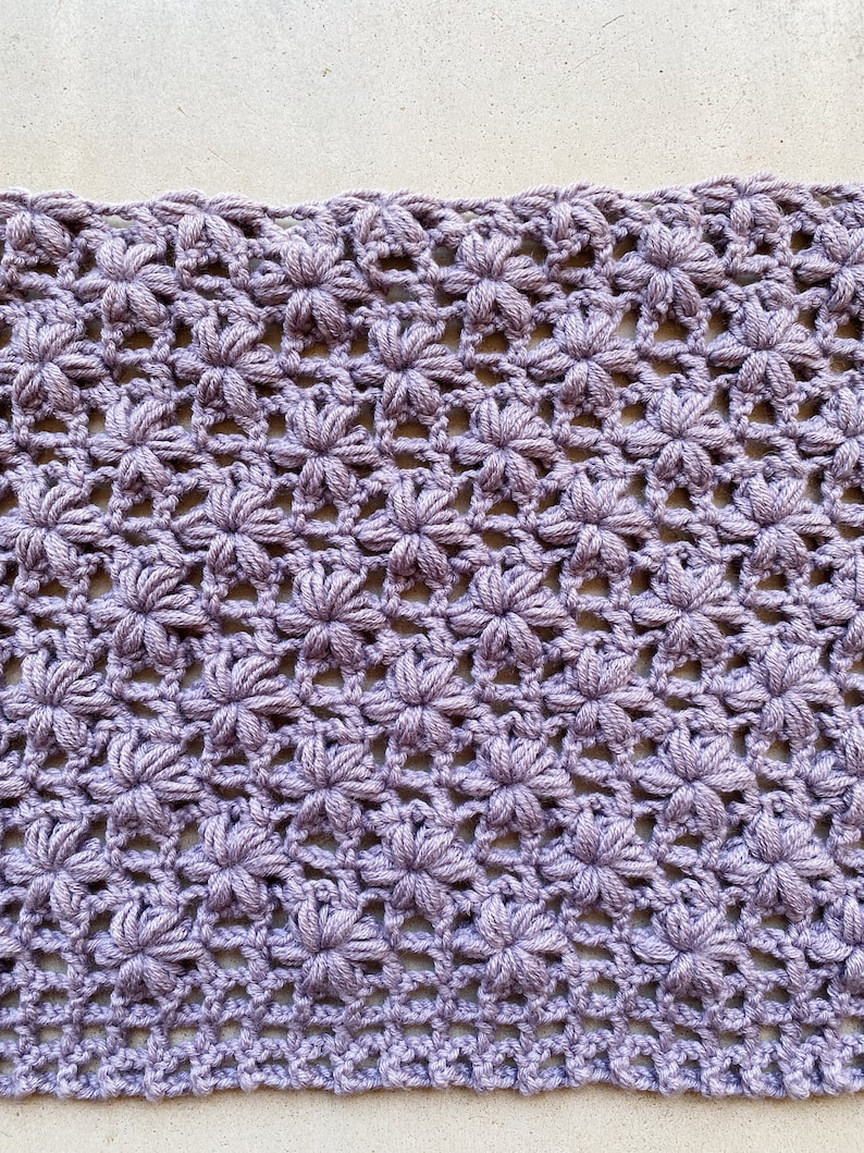 May include: A close-up of a crocheted fabric with a repeating pattern of small flowers. The fabric is a light purple color.
