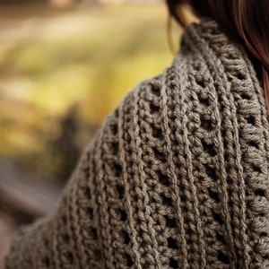 May include: A close-up of a brown crocheted shawl with a textured pattern. The shawl is draped over a person's shoulders.