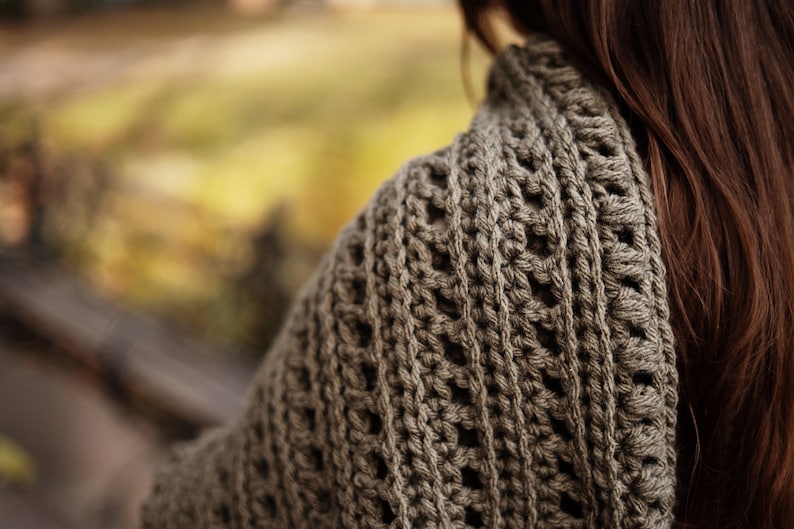 May include: A close-up of a brown crocheted shawl with a textured pattern. The shawl is draped over a person's shoulders.