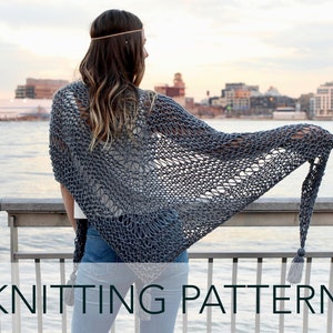 May include: A gray knitted shawl with a tassel hanging from the end. The shawl is draped over a person's shoulders and the person is standing on a pier overlooking a city skyline.