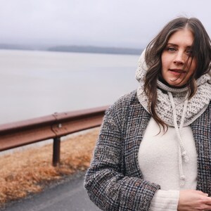 May include: A woman wearing a gray hooded sweater with a white knit scarf, a white sweater, and a gray and black plaid jacket. She is standing in front of a body of water and a metal guardrail.