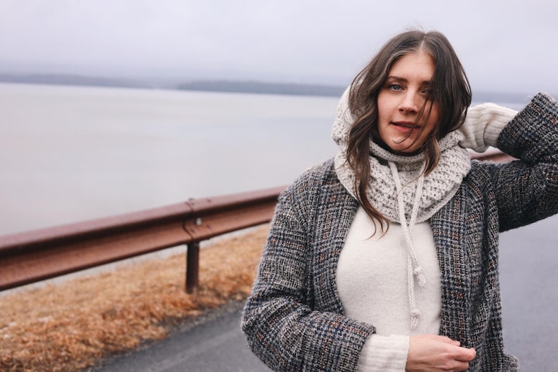 May include: A woman wearing a gray hooded sweater with a white knit scarf, a white sweater, and a gray and black plaid jacket. She is standing in front of a body of water and a metal guardrail.