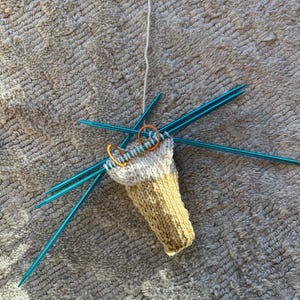 May include: A partially knitted sock in cream and yellow yarn, held by blue knitting needles. Two orange stitch markers are visible. The project is in progress, with a strand of white yarn attached.