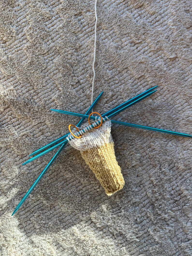 May include: A partially knitted sock in cream and yellow yarn, held by blue knitting needles. Two orange stitch markers are visible. The project is in progress, with a strand of white yarn attached.