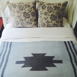 Puede incluir: Una cama blanca con una manta de lana azul claro con un patr&oacute;n geom&eacute;trico gris oscuro. La cama tiene dos almohadas con un patr&oacute;n floral beige y gris.