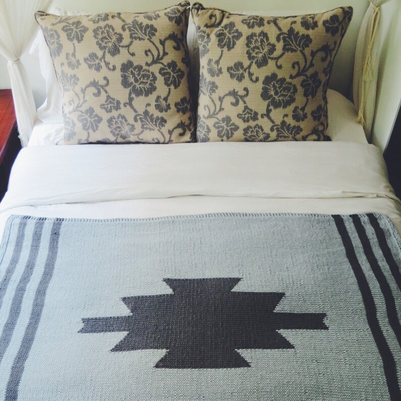 Puede incluir: Una cama blanca con una manta de lana azul claro con un patr&oacute;n geom&eacute;trico gris oscuro. La cama tiene dos almohadas con un patr&oacute;n floral beige y gris.