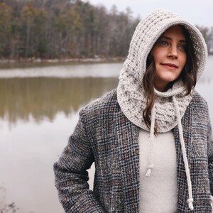 May include: A woman wearing a white crocheted hooded cowl with a drawstring. She is standing in front of a lake with trees in the background.