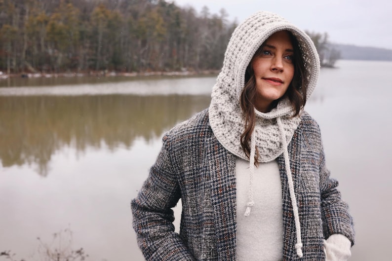 May include: A woman wearing a white crocheted hooded cowl with a drawstring. She is standing in front of a lake with trees in the background.