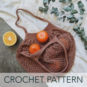 May include: A brown crochet market bag with two oranges inside. The bag has a wide, open top and two long straps. The bag is sitting on a white fabric surface.
