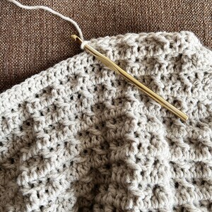 May include: A close-up of a crocheted blanket in a beige color being worked on with a gold crochet hook and white yarn.