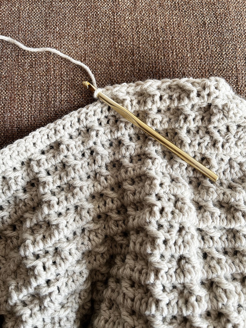 May include: A close-up of a crocheted blanket in a beige color being worked on with a gold crochet hook and white yarn.