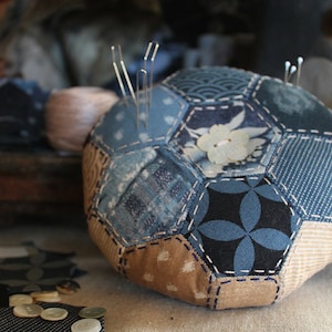Op de afbeelding: Een blauw en bruin patchwork naaldenkussen in de vorm van een bal met spelden die eruit steken. Het naaldenkussen ligt op een tafel met stofresten en knopen die eromheen liggen.