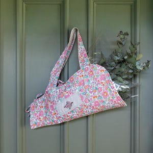 May include: A pink, blue, and white floral tote bag hangs on a green door. The bag features a rectangular patch with a stitched design. The bag's handles are a light pink and white floral pattern. A bundle of greenery is visible.
