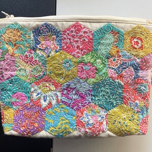 Könnte beinhalten: Eine weiße Stofftasche mit Reißverschluss. Die Tasche ist mit einem Patchwork-Muster aus bunten sechseckigen Stoffstücken verziert. Die Stoffstücke sind mit einem dekorativen Stich zusammengenäht.