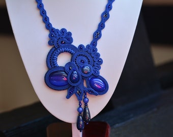Puede incluir: Un collar azul con un gran colgante intrincado hecho de cordón tejido y piedras preciosas azules. El colgante tiene un diseño circular con un círculo más pequeño en el centro. El collar está en un expositor blanco.