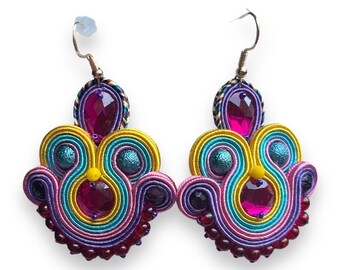 May include: A pair of colorful dangle earrings with a multi-layered design. The earrings feature a mix of pink, yellow, blue, and purple threads, with a large pink crystal in the center of each earring. The earrings are finished with a row of small red beads.