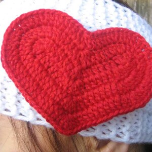 May include: A handmade white crochet headband with a large, vibrant red crochet heart attached. The heart is centrally placed and adds a pop of color to the white headband. The heart is made of a thick yarn.