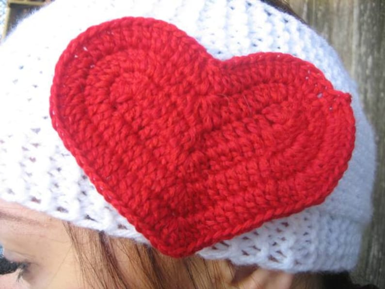 May include: A handmade white crochet headband with a large, vibrant red crochet heart attached. The heart is centrally placed and adds a pop of color to the white headband. The heart is made of a thick yarn.