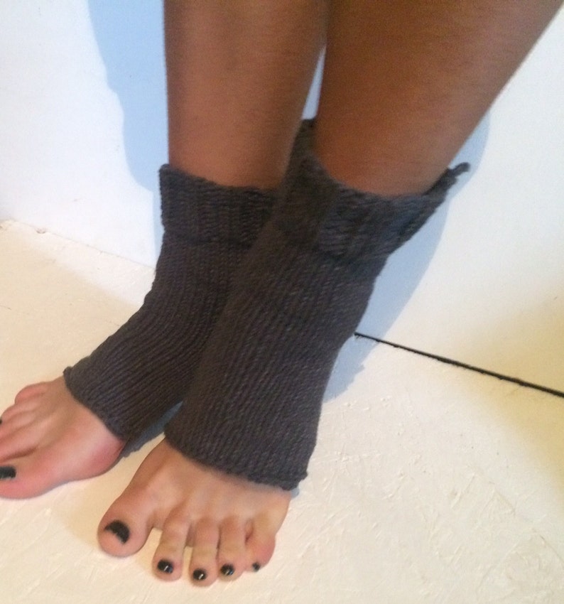 May include: A pair of dark gray knitted leg warmers. The leg warmers are ribbed and extend from the ankles to mid-calf. The image shows the leg warmers on a person's legs, with the feet visible.