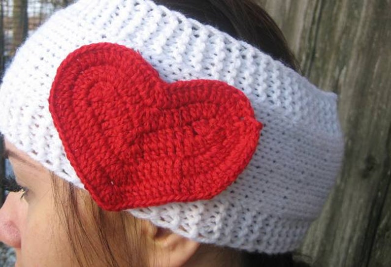 May include: A white crocheted headband with a large, red heart applique. The heart is also crocheted and is centered on the headband. The headband is a winter accessory, perfect for Valentine's Day or everyday wear.