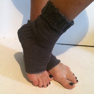 May include: Dark gray knitted foot warmers. The warmers are made of a thick, textured yarn and cover the foot and ankle. The top of the warmer is folded over. The background is a white surface.