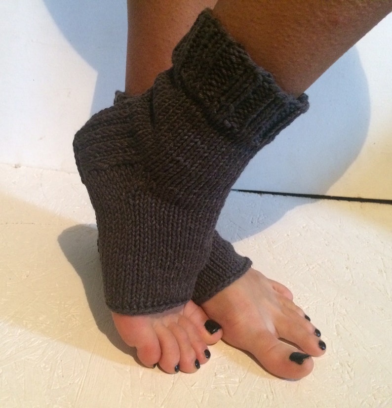 May include: Dark gray knitted foot warmers. The warmers are made of a thick, textured yarn and cover the foot and ankle. The top of the warmer is folded over. The background is a white surface.