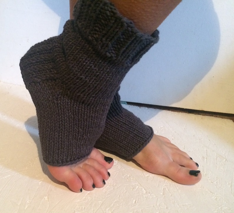May include: Dark gray knitted leg warmers. The leg warmers are made of a thick, textured yarn and have a ribbed cuff at the top. The foot portion is open, and the leg warmers are designed to be worn over the foot and ankle.