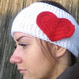 May include: A white knitted headband with a large red crocheted heart attached to the side. The headband is made of a thick, textured yarn. The heart is a vibrant red color.