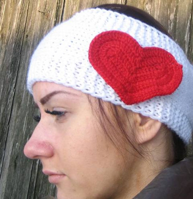 May include: A white knitted headband with a large red crocheted heart attached to the side. The headband is made of a thick, textured yarn. The heart is a vibrant red color.