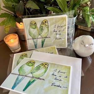 May include: A set of greeting cards featuring a watercolor illustration of two green budgies perched on a branch. The text reads, A friend loves at all times. Proverbs 17:17. The cards are packaged in clear plastic sleeves with white envelopes.