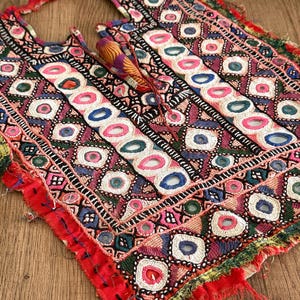 Op de afbeelding: Een rechthoekig textielpaneel met ingewikkeld handborduurwerk met geometrische patronen, kleine ronde spiegels en levendige draadkleuren waaronder rood, roze, blauw en groen. De randen zijn gerafeld met losse draden en stofresten.
