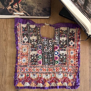 Op de afbeelding: Een paars stoffen paneel met ingewikkeld handgestikt borduurwerk in zwarte, witte, oranje en groene draden. Het textiel is versierd met kleine ronde spiegeltjes en geometrische patronen, met een centrale halsopening en gerafelde randen.
