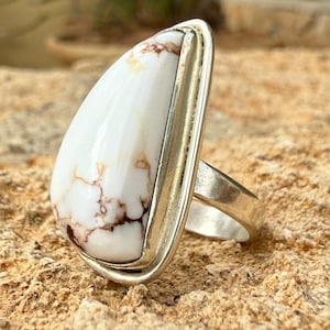 Op de afbeelding: Een zilveren ring met een grote, druppelvormige witte steen met bruine adering. De steen is gezet in een zilveren bezel en heeft een gepolijste afwerking. De ringband is van gepolijst zilver.