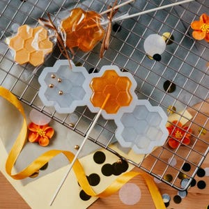 Può includere: Lecca lecca a forma di favo e uno stampo in silicone su una griglia di raffreddamento in metallo. I lecca lecca sono ambrati e gialli, alcuni avvolti in plastica trasparente. Fiori arancioni, nastro dorato e coriandoli decorano la scena.