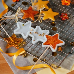 May include: A close-up of star-shaped lollipops on a wire rack. The lollipops are orange, yellow, and white with glitter. The lollipops are on a white surface with a yellow ribbon.