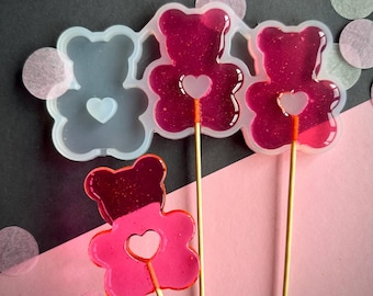 May include: A close-up shot features three red bear-shaped lollipops on wooden sticks, each with a heart-shaped center. A clear silicone mold with a bear shape is also visible. The lollipops are on a pink and black background with confetti.