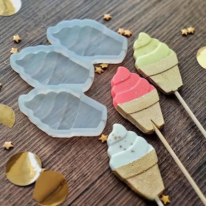 Puede incluir: Una vista aérea muestra tres moldes en forma de cono de helado y tres helados decorados en palitos. Los helados tienen helado suave rosa, verde y azul con conos dorados. Confeti dorado y chispas en forma de estrella están esparcidos.