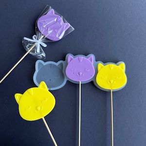 May include: A collection of cat-shaped lollipops and molds in various colors. One lollipop is purple and wrapped in clear cellophane. The molds are gray, purple, and yellow. The lollipops and molds are on wooden sticks.