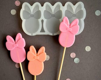 May include: A silicone mould with three bow-shaped cavities, next to three bow-shaped lollipops in pink and orange hues. The lollipops are on wooden sticks, set against a dark grey background.